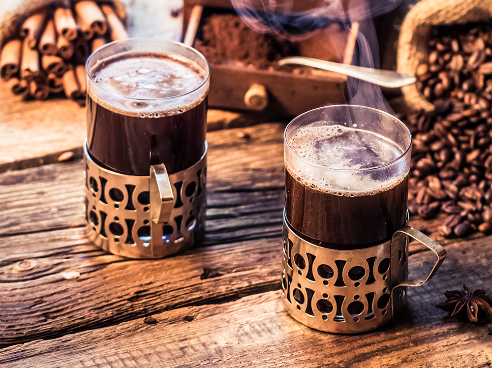 Two steaming cups of His Word Coffee in vintage holders on a rustic wooden table, surrounded by fresh coffee beans and cinnamon sticks, evoking warmth and artisanal quality.