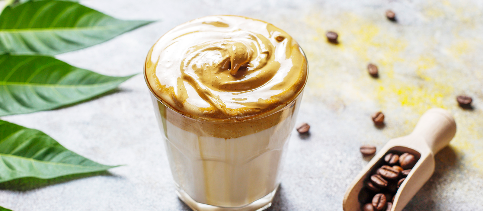 Whipped Dalgona-style His Word Coffee in a glass over iced milk, surrounded by coffee beans and tropical leaves—celebrating creative brewing and faith-filled coffee moments.